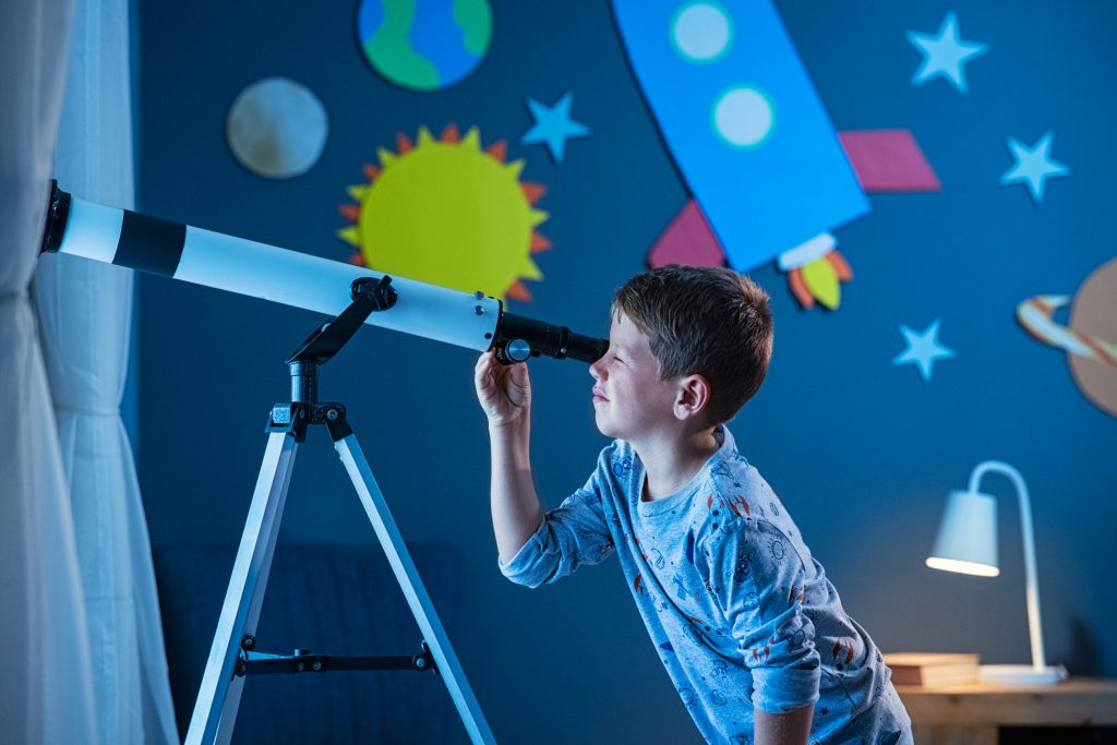 Curious boy using telescope to explore moon surface at night in his bedroom. Young child using telescope to see remote galaxy from room with decorated wall with rocket, planets, stars and cardboard solar system. Passionate kid in pajamas looking at stars using telescope, astronomy and stargazing concept.